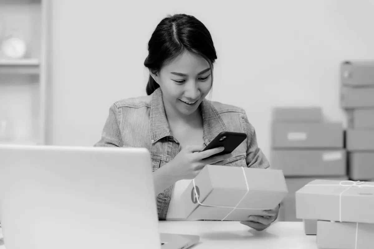 A woman smiling at her phone while packing boxes for shipping