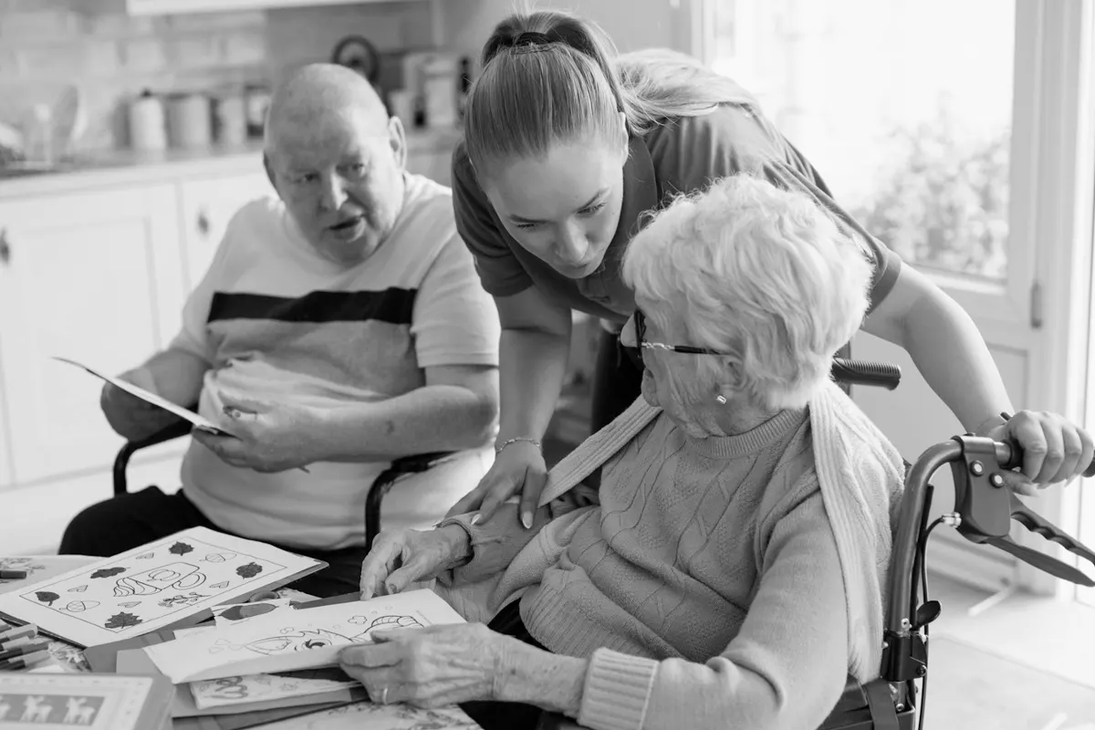 A caregiver helping two older residents with an activity at a table